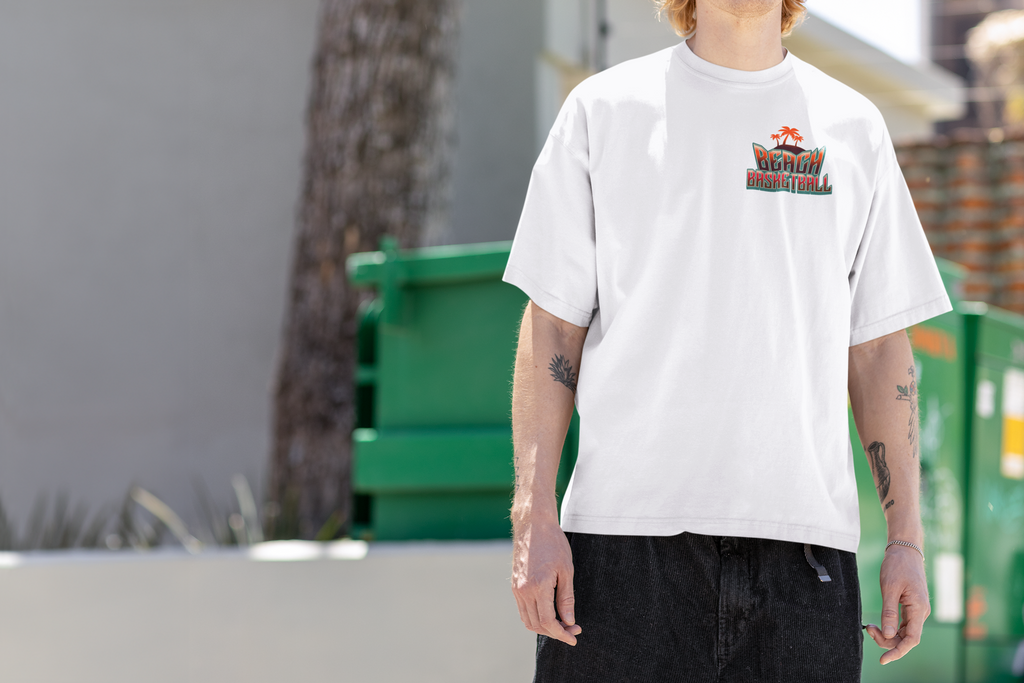 Kleiner "Beach Basketball" Retro-Print mit Palmen in Orange und Türkis auf der linken Brust eines Mockup mit Model eines weißen Oversized T-Shirts. 100% Baumwolle