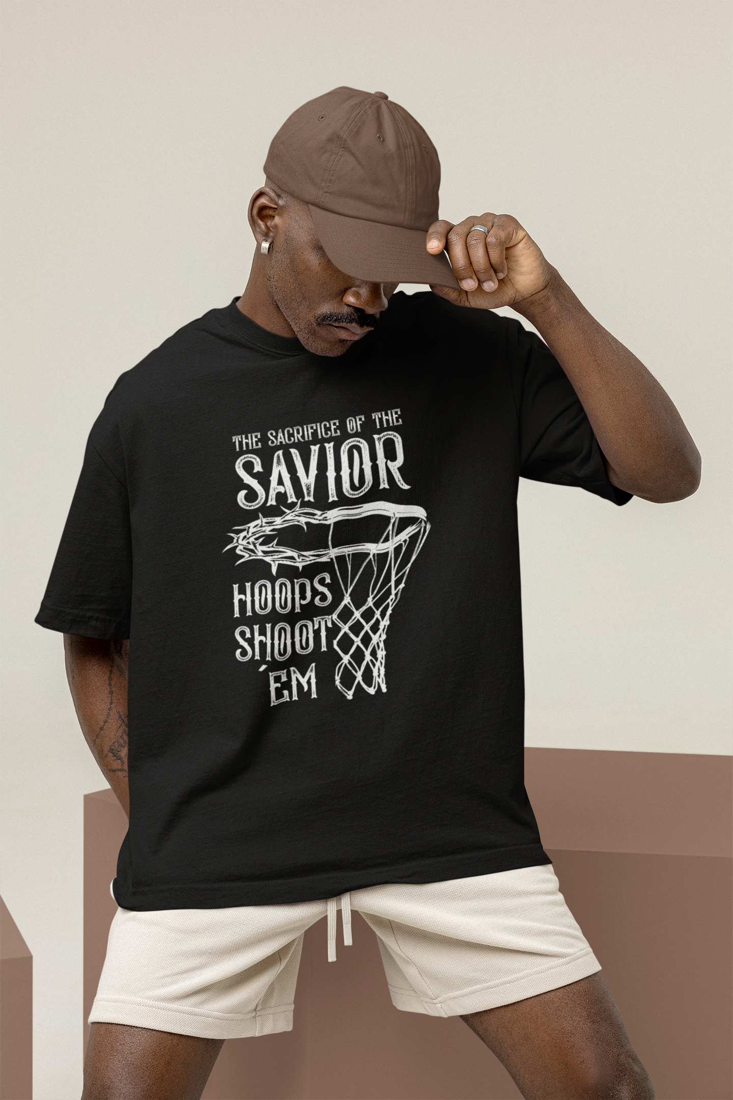 Mockup eines Mannes mit Schwarzen Oversized T-Shirt mit einem weißen Print, der einen Basketballkorb zeigt, dessen Ring eine Dornenkrone ist. Darüber steht der Text „THE SACRIFICE OF THE SAVIOR“ und darunter „HOOPS SHOOT EM“. 100% Baumwolle.