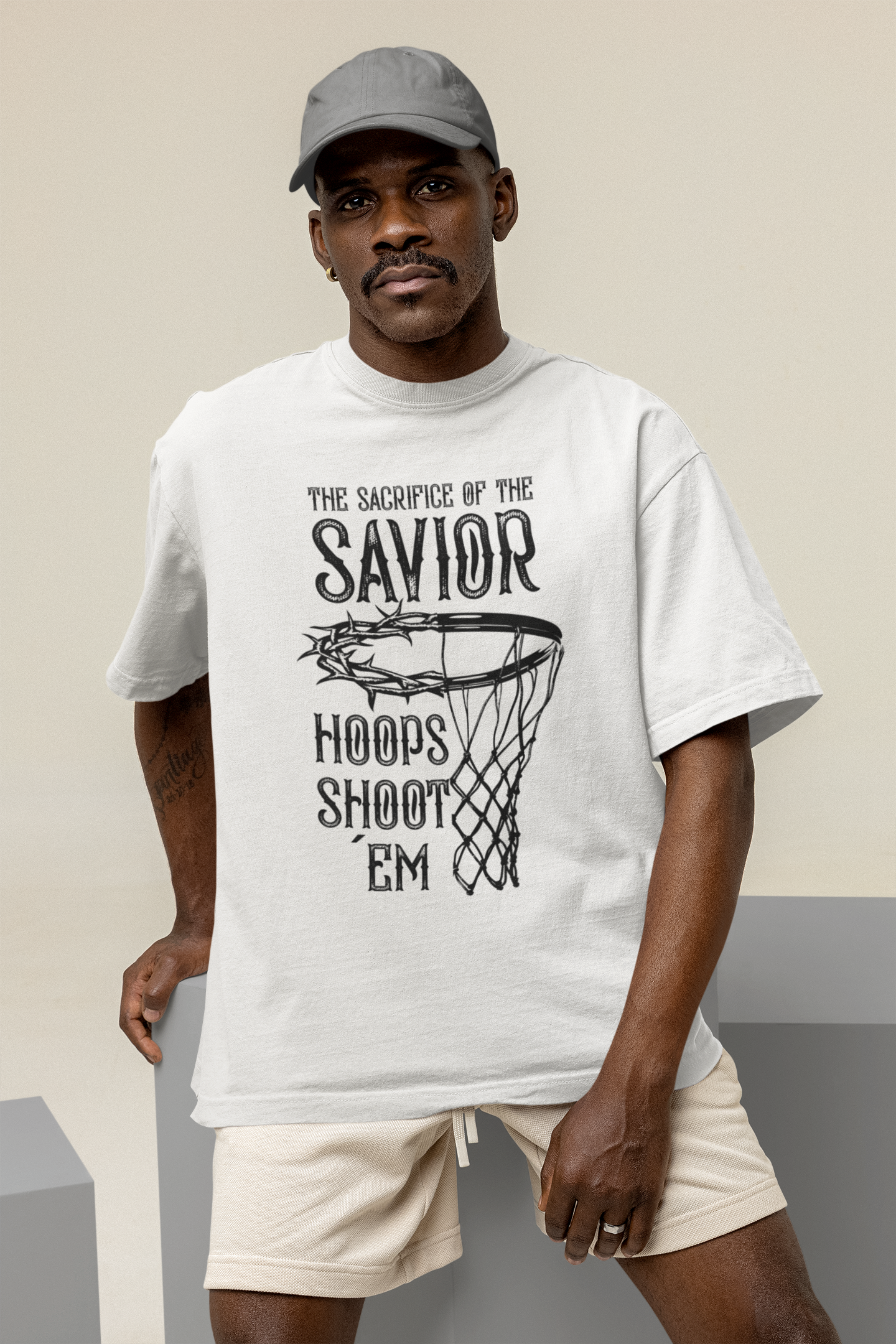Mockup mit einen Mann Weißen Oversized T-Shirt mit einem weißen Print, der einen Basketballkorb zeigt, dessen Ring eine Dornenkrone ist. Darüber steht der Text „THE SACRIFICE OF THE SAVIOR“ und darunter „HOOPS SHOOT EM“. 100% Baumwolle.