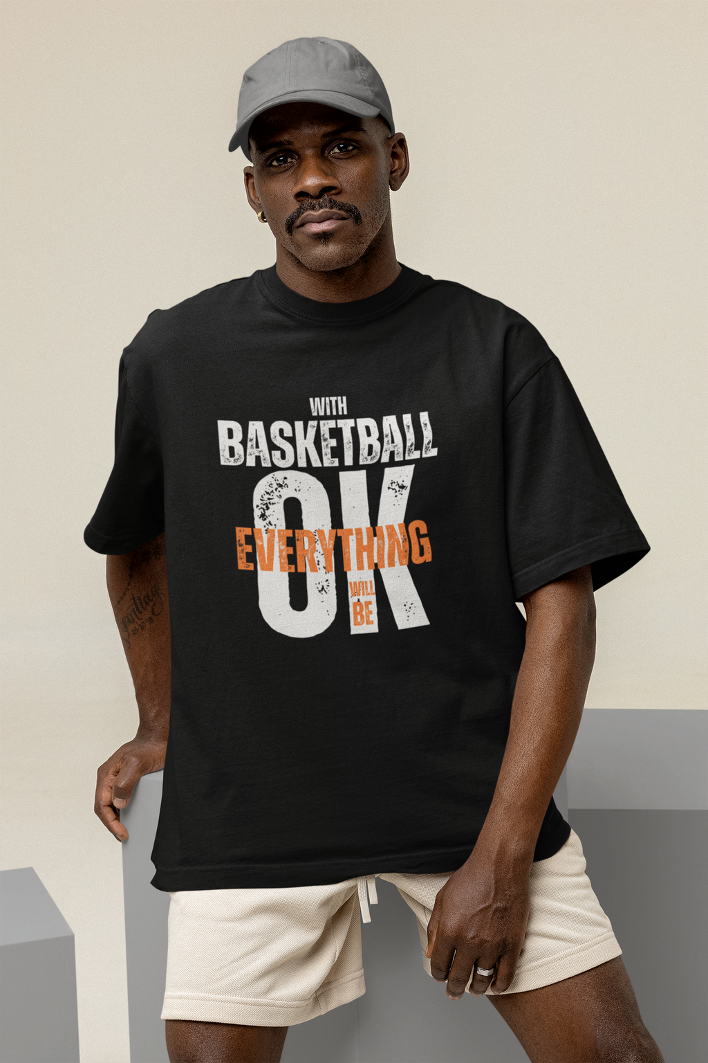 Männliches Model trägt Schwarzes Oversized T-Shirt aus 100% Baumwolle mit dem Aufdruck "WITH BASKETBALL EVERYTHING WILL BE OK". Der Text ist in Weiß und Orange gehalten, wobei "EVERYTHING" und "BE" in Orange hervorgehoben sind.