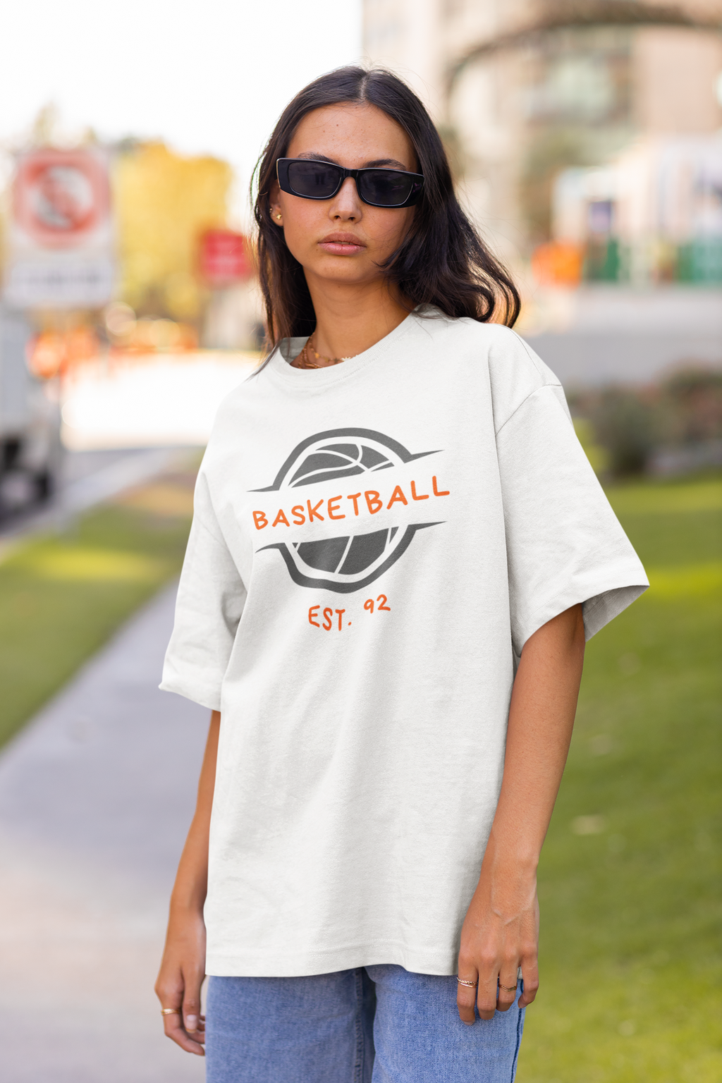 Mockup mit Model Weißes Oversized T-Shirt mit einem Print eines grauen Basketball-Emblems und dem orangefarbenen, stilisierten Text „BASKETBALL“ und „EST. 92“. 100% Baumwolle.