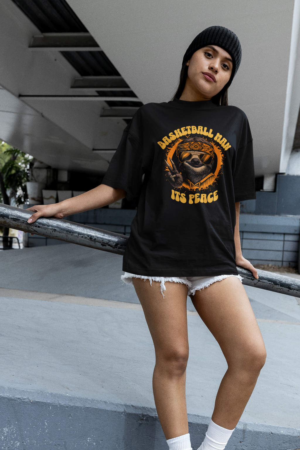 Mockup mit Model Schwarzes Oversized  Basketball T-Shirt mit einem 70er-Jahre-Retro-Print eines coolen Faultiers (Sloth) mit Sonnenbrille und Bandana, das ein Peace-Zeichen macht. Der Text lautet „BASKETBALL MAN“ und „ITS PEACE“ in Orange und Gelb. 100% Baumwolle.